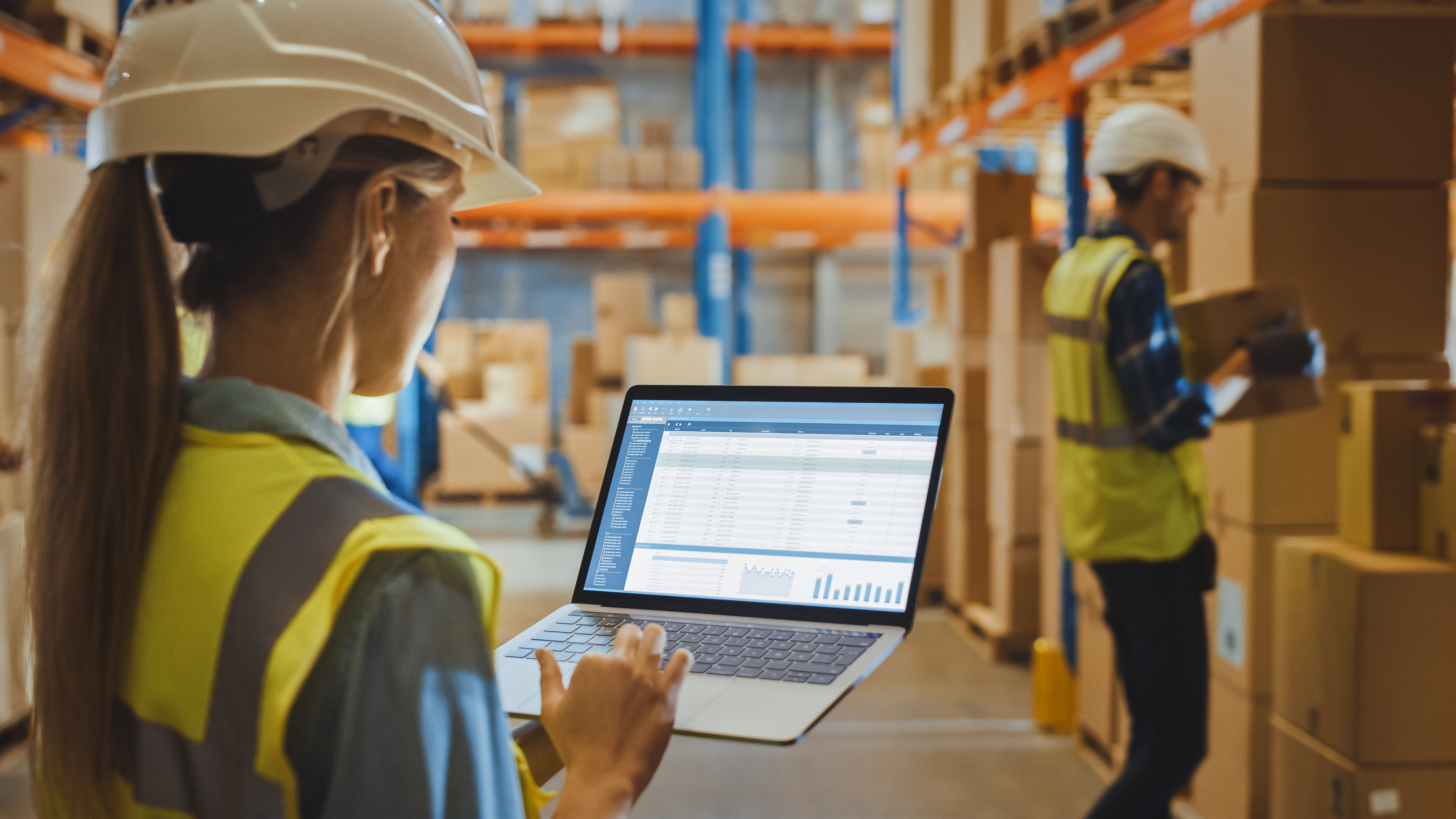 Vrouwelijke magazijnmedewerker met veiligheidshelm houdt een laptop vast en bekijkt een rapport over Warehouse Profitability.
